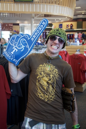 A person in a store wearing a brown graphic t-shirt, plaid shorts, sunglasses, and a knit hat. They are holding a large blue foam hand with 'Rangers' on it, and have a wide grin. The store appears to have sports merchandise, visible in the background.