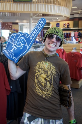 A person in a store wearing a brown graphic t-shirt, plaid shorts, sunglasses, and a knit hat. They are holding a large blue foam hand with 'Rangers' on it, and have a wide grin. The store appears to have sports merchandise, visible in the background.