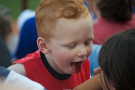 A young child with red hair and a red shirt is leaning forward with their tongue out, appearing playful. The background includes several other people suggesting a social environment.