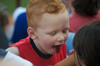 A young child with red hair and a red shirt is leaning forward with their tongue out, appearing playful. The background includes several other people suggesting a social environment.