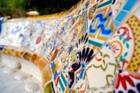A detailed mosaic with an array of colorful, geometric, and floral patterns. The tiles are intricately arranged, showcasing a blend of shapes and vibrant colors like blue, orange, and yellow. The artwork seems to cover a large, curved surface, possibly part of a bench or wall.