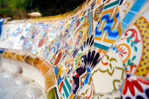 A detailed mosaic with an array of colorful, geometric, and floral patterns. The tiles are intricately arranged, showcasing a blend of shapes and vibrant colors like blue, orange, and yellow. The artwork seems to cover a large, curved surface, possibly part of a bench or wall.