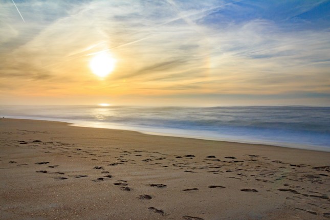 A peaceful beach scene with gentle ocean waves lapping onto the sandy shore, illuminated by a warm, glowing sunset. The sky is a blend of soft orange and blue hues with scattered clouds, adding texture to the horizon.