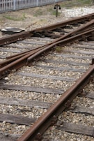 Railway tracks converge and diverge, surrounded by gravel and wooden sleepers. A metal signal or switch device is visible in the background, with a fence bordering the area.