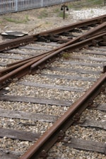 Railway tracks converge and diverge, surrounded by gravel and wooden sleepers. A metal signal or switch device is visible in the background, with a fence bordering the area.