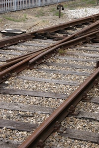 Railway tracks converge and diverge, surrounded by gravel and wooden sleepers. A metal signal or switch device is visible in the background, with a fence bordering the area.
