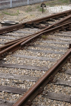 Railway tracks converge and diverge, surrounded by gravel and wooden sleepers. A metal signal or switch device is visible in the background, with a fence bordering the area.