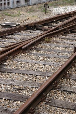 Railway tracks converge and diverge, surrounded by gravel and wooden sleepers. A metal signal or switch device is visible in the background, with a fence bordering the area.