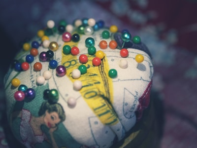 A cushion is adorned with colorful push pins, each having a small round head. The cushion features printed images including a vintage-style illustration of a woman and measuring tape designs.
