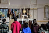A group of people are gathered in front of an ornate altar in a church. The altar is decorated with numerous lit candles, a golden monstrance, flowers, and a white cloth. There's a statue on the right side and stained glass windows in the background.