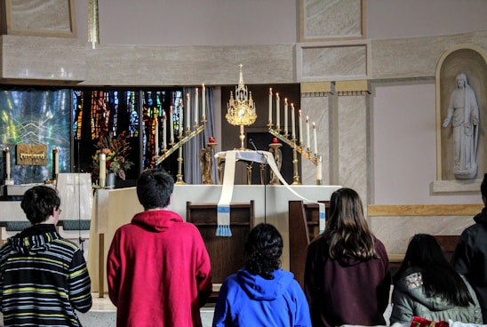 A group of people are gathered in front of an ornate altar in a church. The altar is decorated with numerous lit candles, a golden monstrance, flowers, and a white cloth. There's a statue on the right side and stained glass windows in the background.