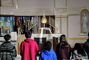 A group of people are gathered in front of an ornate altar in a church. The altar is decorated with numerous lit candles, a golden monstrance, flowers, and a white cloth. There's a statue on the right side and stained glass windows in the background.