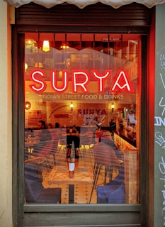 A warm and inviting view of a cozy restaurant interior with a prominent neon sign reading 'Surya' above the text 'Indian Street Food & Drinks'. The inside features a rustic decor with wooden tables and a mixture of chairs. Dim lighting and pendant lamps create a relaxed atmosphere, while reflections in the window hint at street art and an urban setting outside.