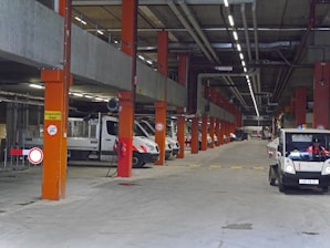 A large underground parking or storage area featuring several industrial vehicles parked in rows. The space is supported by orange structural columns and has overhead piping. Signs are visible, including one indicating a speed limit and others for traffic instructions. The lighting is artificial, coming from overhead strip lights.