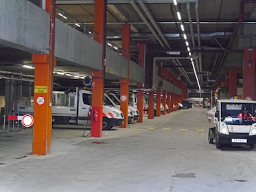 A large underground parking or storage area featuring several industrial vehicles parked in rows. The space is supported by orange structural columns and has overhead piping. Signs are visible, including one indicating a speed limit and others for traffic instructions. The lighting is artificial, coming from overhead strip lights.