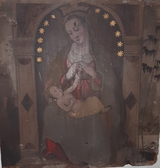 A painting depicts a seated woman adorned with a crown of stars, cradling a child in her arms. The background features architectural elements and muted, earthy tones. The woman is dressed in a robe with a veil, creating a serene, contemplative atmosphere.