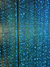 A visual display of numerous vertical strands of LED lights emitting a vibrant glow in shades of blue and green. The lights are densely arranged, creating an intricate and nearly hypnotic pattern with subtle reflections and refractions that enhance the immersive effect.