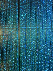 A visual display of numerous vertical strands of LED lights emitting a vibrant glow in shades of blue and green. The lights are densely arranged, creating an intricate and nearly hypnotic pattern with subtle reflections and refractions that enhance the immersive effect.