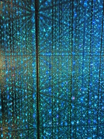 A visual display of numerous vertical strands of LED lights emitting a vibrant glow in shades of blue and green. The lights are densely arranged, creating an intricate and nearly hypnotic pattern with subtle reflections and refractions that enhance the immersive effect.