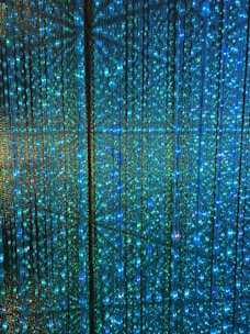 A visual display of numerous vertical strands of LED lights emitting a vibrant glow in shades of blue and green. The lights are densely arranged, creating an intricate and nearly hypnotic pattern with subtle reflections and refractions that enhance the immersive effect.
