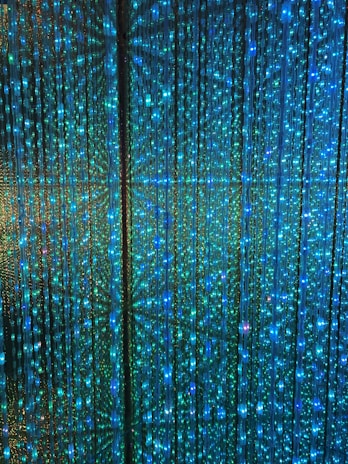 A visual display of numerous vertical strands of LED lights emitting a vibrant glow in shades of blue and green. The lights are densely arranged, creating an intricate and nearly hypnotic pattern with subtle reflections and refractions that enhance the immersive effect.