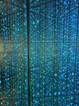 A visual display of numerous vertical strands of LED lights emitting a vibrant glow in shades of blue and green. The lights are densely arranged, creating an intricate and nearly hypnotic pattern with subtle reflections and refractions that enhance the immersive effect.