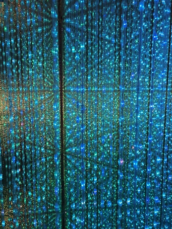 A visual display of numerous vertical strands of LED lights emitting a vibrant glow in shades of blue and green. The lights are densely arranged, creating an intricate and nearly hypnotic pattern with subtle reflections and refractions that enhance the immersive effect.