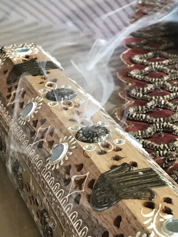 A detailed wooden incense holder with intricate carvings and designs, emitting streams of smoke. Decorative patterns are etched on the surface, and there is an ornate fabric visible in the background.