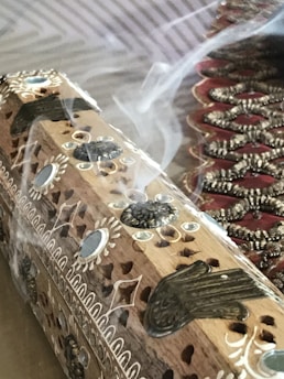 A detailed wooden incense holder with intricate carvings and designs, emitting streams of smoke. Decorative patterns are etched on the surface, and there is an ornate fabric visible in the background.