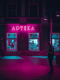 A neon-lit pharmacy with a vibrant pink sign illuminates the surrounding area. The windows display various items with a turquoise glow, contrasting with the pink light. A silhouette of a person is visible on the sidewalk, engrossed in their phone.