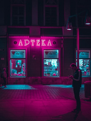 A neon-lit pharmacy with a vibrant pink sign illuminates the surrounding area. The windows display various items with a turquoise glow, contrasting with the pink light. A silhouette of a person is visible on the sidewalk, engrossed in their phone.