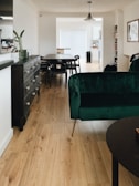 A modern and stylish interior featuring a wooden floor, a dark green velvet sofa, and a dark wooden table with matching chairs. The room is well-lit with natural light coming from a large window adorned with white blinds. A dark wooden sideboard with drawers is placed against the wall, and a black pendant light hangs from the ceiling. The walls are decorated with a few artworks and shelves containing books.