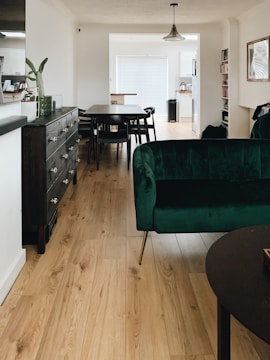 A modern and stylish interior featuring a wooden floor, a dark green velvet sofa, and a dark wooden table with matching chairs. The room is well-lit with natural light coming from a large window adorned with white blinds. A dark wooden sideboard with drawers is placed against the wall, and a black pendant light hangs from the ceiling. The walls are decorated with a few artworks and shelves containing books.