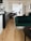 A modern and stylish interior featuring a wooden floor, a dark green velvet sofa, and a dark wooden table with matching chairs. The room is well-lit with natural light coming from a large window adorned with white blinds. A dark wooden sideboard with drawers is placed against the wall, and a black pendant light hangs from the ceiling. The walls are decorated with a few artworks and shelves containing books.