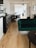A modern and stylish interior featuring a wooden floor, a dark green velvet sofa, and a dark wooden table with matching chairs. The room is well-lit with natural light coming from a large window adorned with white blinds. A dark wooden sideboard with drawers is placed against the wall, and a black pendant light hangs from the ceiling. The walls are decorated with a few artworks and shelves containing books.