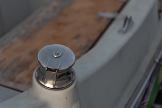 A close-up view of a metallic boat cleat mounted on the edge of a boat. The cleat has a polished finish and is attached to an off-white surface. The background reveals a blurred view of the boat's deck, made of wood, with some hardware slightly visible.