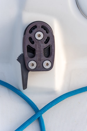 A close-up view of a pulley system mounted on a flat surface, featuring two visible screws and the brand name 'Harken' engraved. A blue rope is seen passing beneath the pulley.