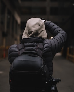 A person wearing a hooded jacket and backpack is standing in a dimly lit alleyway. The scene conveys a sense of solitude and urban exploration.