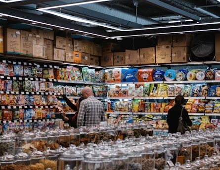 A busy grocery store aisle filled with shelves stocked with a variety of snacks and packaged goods. People browse the selection, with numerous jars containing various dried foods in the foreground. Cardboard boxes are stacked above the shelves, and fluorescent lighting illuminates the space.