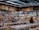 A busy grocery store aisle filled with shelves stocked with a variety of snacks and packaged goods. People browse the selection, with numerous jars containing various dried foods in the foreground. Cardboard boxes are stacked above the shelves, and fluorescent lighting illuminates the space.