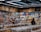 A busy grocery store aisle filled with shelves stocked with a variety of snacks and packaged goods. People browse the selection, with numerous jars containing various dried foods in the foreground. Cardboard boxes are stacked above the shelves, and fluorescent lighting illuminates the space.