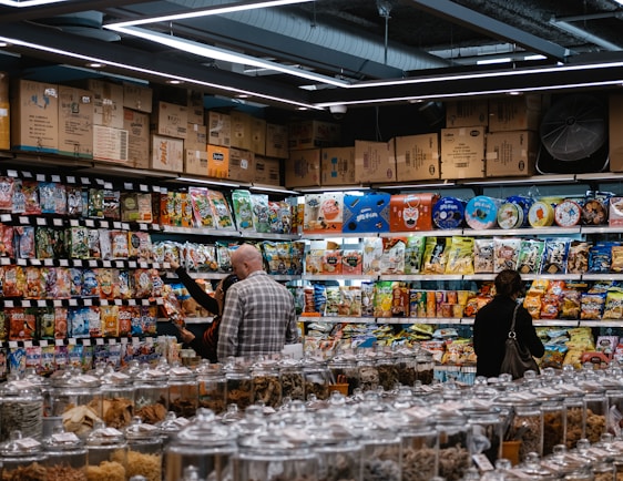 A busy grocery store aisle filled with shelves stocked with a variety of snacks and packaged goods. People browse the selection, with numerous jars containing various dried foods in the foreground. Cardboard boxes are stacked above the shelves, and fluorescent lighting illuminates the space.