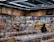A busy grocery store aisle filled with shelves stocked with a variety of snacks and packaged goods. People browse the selection, with numerous jars containing various dried foods in the foreground. Cardboard boxes are stacked above the shelves, and fluorescent lighting illuminates the space.