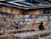 A busy grocery store aisle filled with shelves stocked with a variety of snacks and packaged goods. People browse the selection, with numerous jars containing various dried foods in the foreground. Cardboard boxes are stacked above the shelves, and fluorescent lighting illuminates the space.