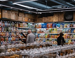 A busy grocery store aisle filled with shelves stocked with a variety of snacks and packaged goods. People browse the selection, with numerous jars containing various dried foods in the foreground. Cardboard boxes are stacked above the shelves, and fluorescent lighting illuminates the space.