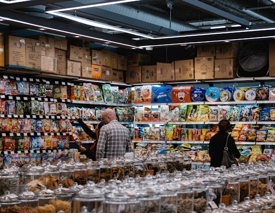 A busy grocery store aisle filled with shelves stocked with a variety of snacks and packaged goods. People browse the selection, with numerous jars containing various dried foods in the foreground. Cardboard boxes are stacked above the shelves, and fluorescent lighting illuminates the space.