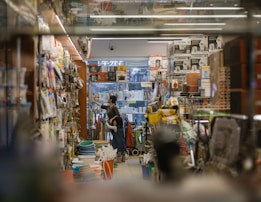 A cluttered store aisle filled with various household items and appliances. A person and a child are browsing through the products, with shelves lined with goods on either side. The environment appears busy and commercial.