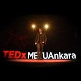 A speaker is on stage at a TEDx event, with large illuminated letters spelling out TEDxMETUAnkara behind him. The stage is dimly lit with spotlights above the speaker, creating a professional and engaging atmosphere.