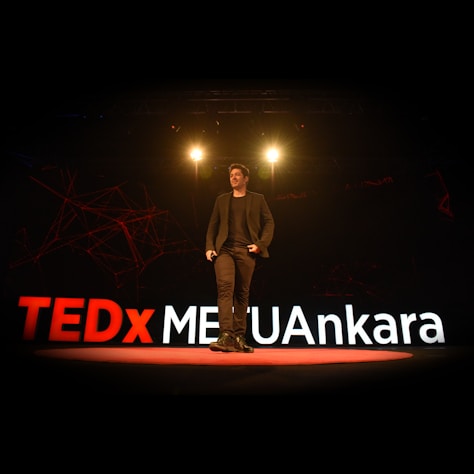 A speaker is on stage at a TEDx event, with large illuminated letters spelling out TEDxMETUAnkara behind him. The stage is dimly lit with spotlights above the speaker, creating a professional and engaging atmosphere.