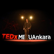 A speaker is on stage at a TEDx event, with large illuminated letters spelling out TEDxMETUAnkara behind him. The stage is dimly lit with spotlights above the speaker, creating a professional and engaging atmosphere.